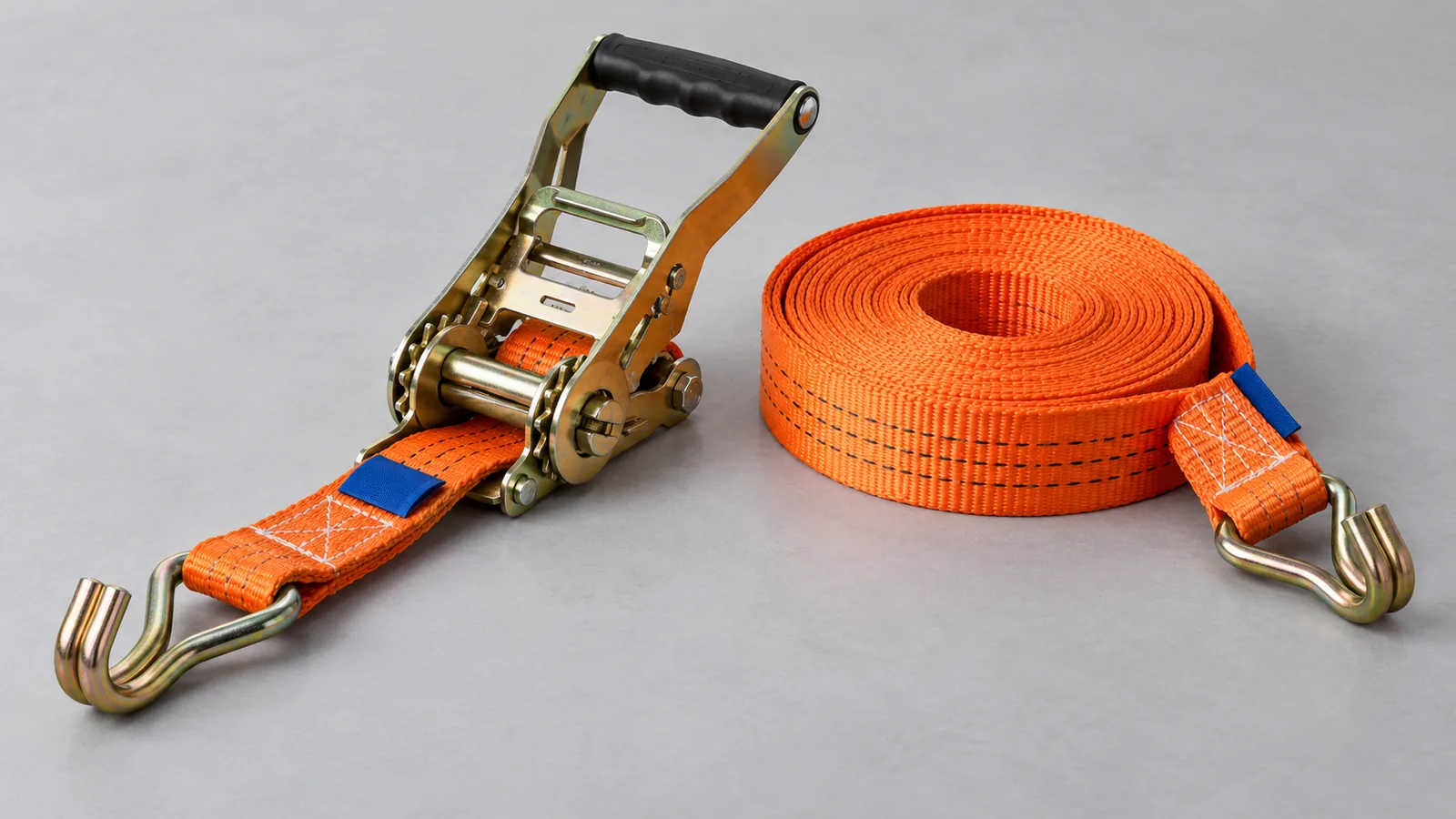 Orange ratchet tie-down webbing with a tensioner and double J hooks on a grey tabletop